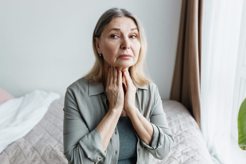 Indoor portrait of worried senior woman siting on bed in bedroom examining her thyroid, touching her throat, looking aside with anxiety, trying to find symptoms of severe endocrine disease